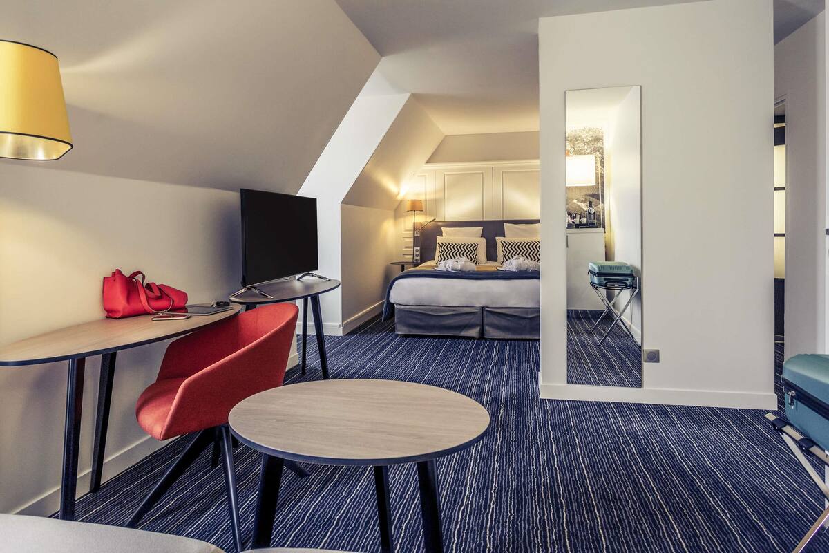 Ein Hotelzimmer mit Schreibtisch, einem roten Sessel, einem Bett, einem Fernseher und einem durchgegangenen Tür sichtbar erkennbaren Badezimmer.