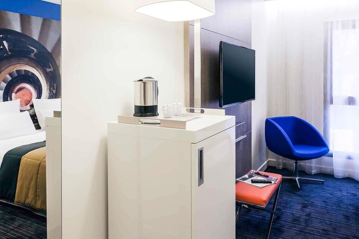 Une chambre d’hôtel équipée d’une kitchenette, d’un téléviseur à écran plat, d’un fauteuil bleu et d’une fenêtre avec des rideaux.