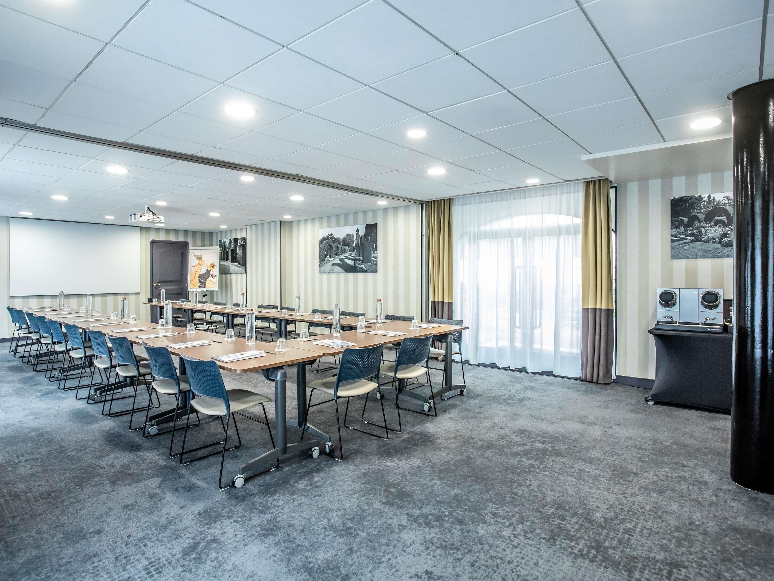 A conference room with long tables, chairs, a projector screen, and a large window with curtains.