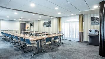 A conference room with long tables, chairs, a projector screen, and a large window with curtains.