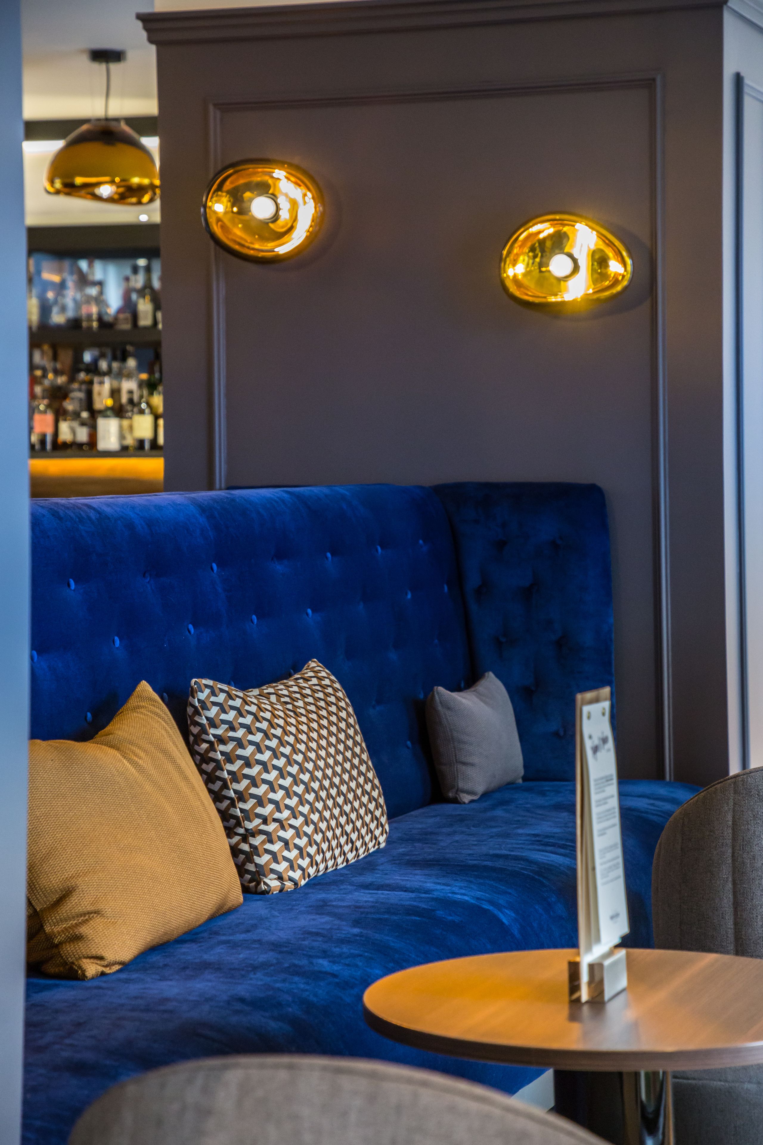 Um sofá azul com almofadas estampadas, uma mesa redonda de madeira e duas luminárias pendentes.