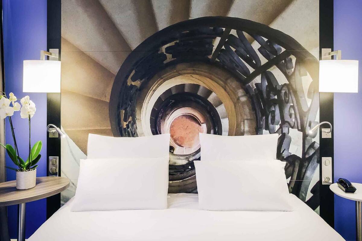 Ein Hotelzimmer mit einem Bett, zwei Nachttischlampen, einem Nachttisch mit Telefon und einer Wand mit einem großen abstrakten Gemälde.