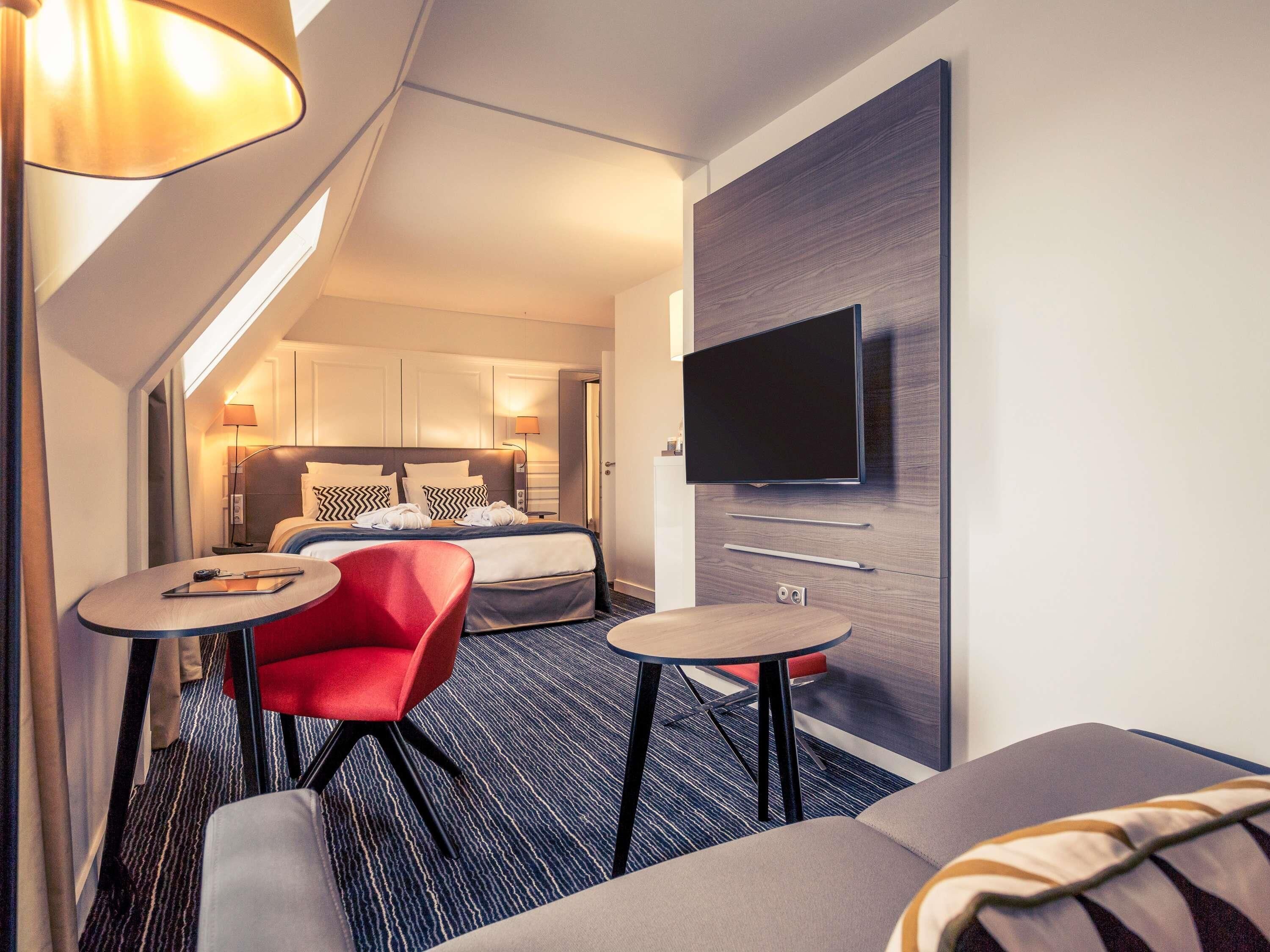 Habitación de hotel con cama, televisor montado en la pared, dos mesitas, una silla roja y un sofá.