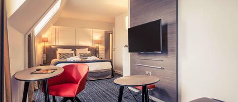 Habitación de hotel con cama, televisor montado en la pared, dos mesitas, una silla roja y un sofá.