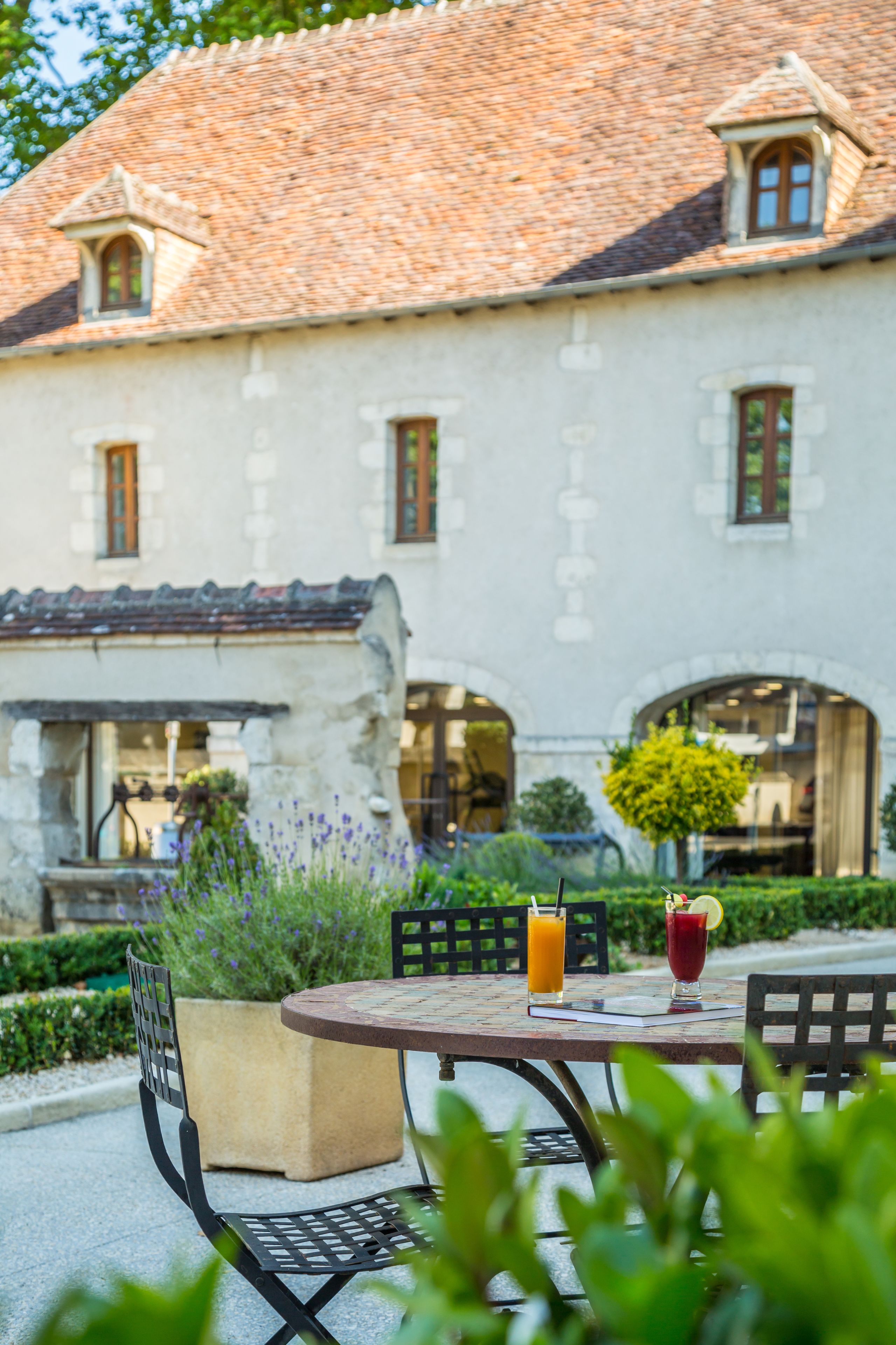 An outdoor dining area with a table and chairs, a building with a tiled roof, and a garden with purple flowers.