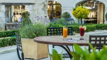 An outdoor dining area with a table and chairs, a building with a tiled roof, and a garden with purple flowers.