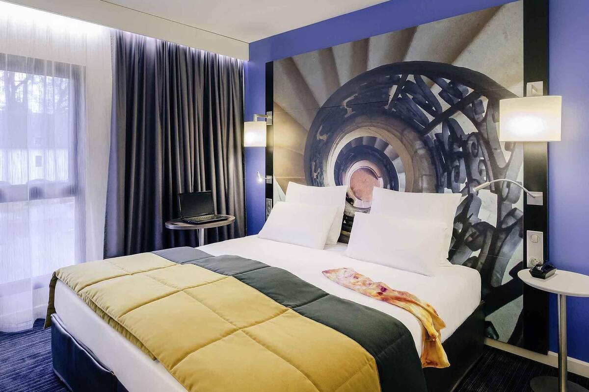 Ein Hotelzimmer mit einem Bett, einem Schreibtisch, einem Sessel und einer Lampe. An der Wand hängt ein großes, abstraktes Gemälde.