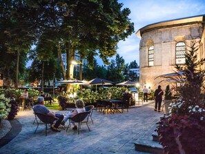 Un espace extérieur aménagé avec des tables et des chaises, entouré d’arbres et bordé d’un bâtiment en pierre.