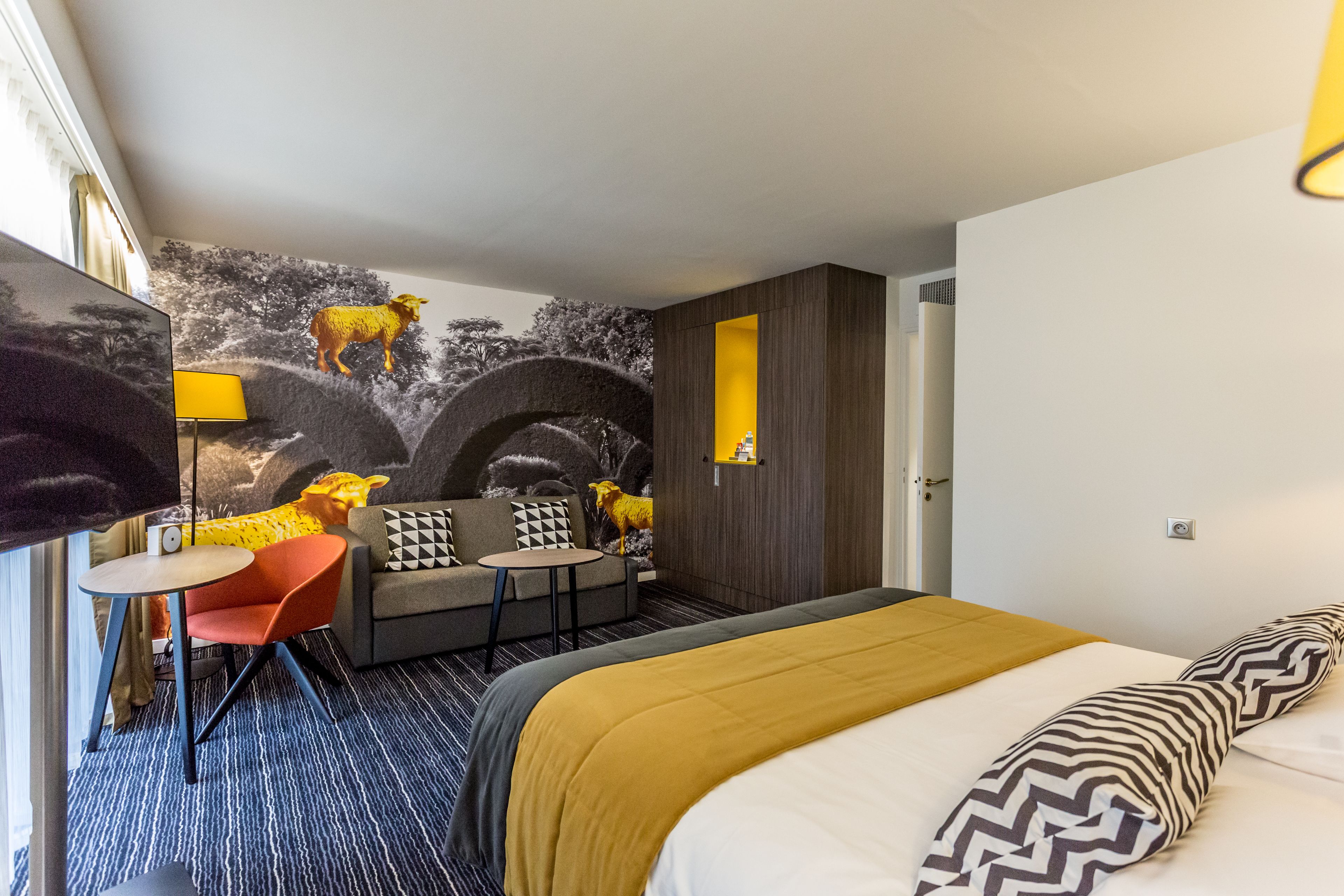 A hotel room with a bed, a sofa, a small table, a chair, and a wall mural.