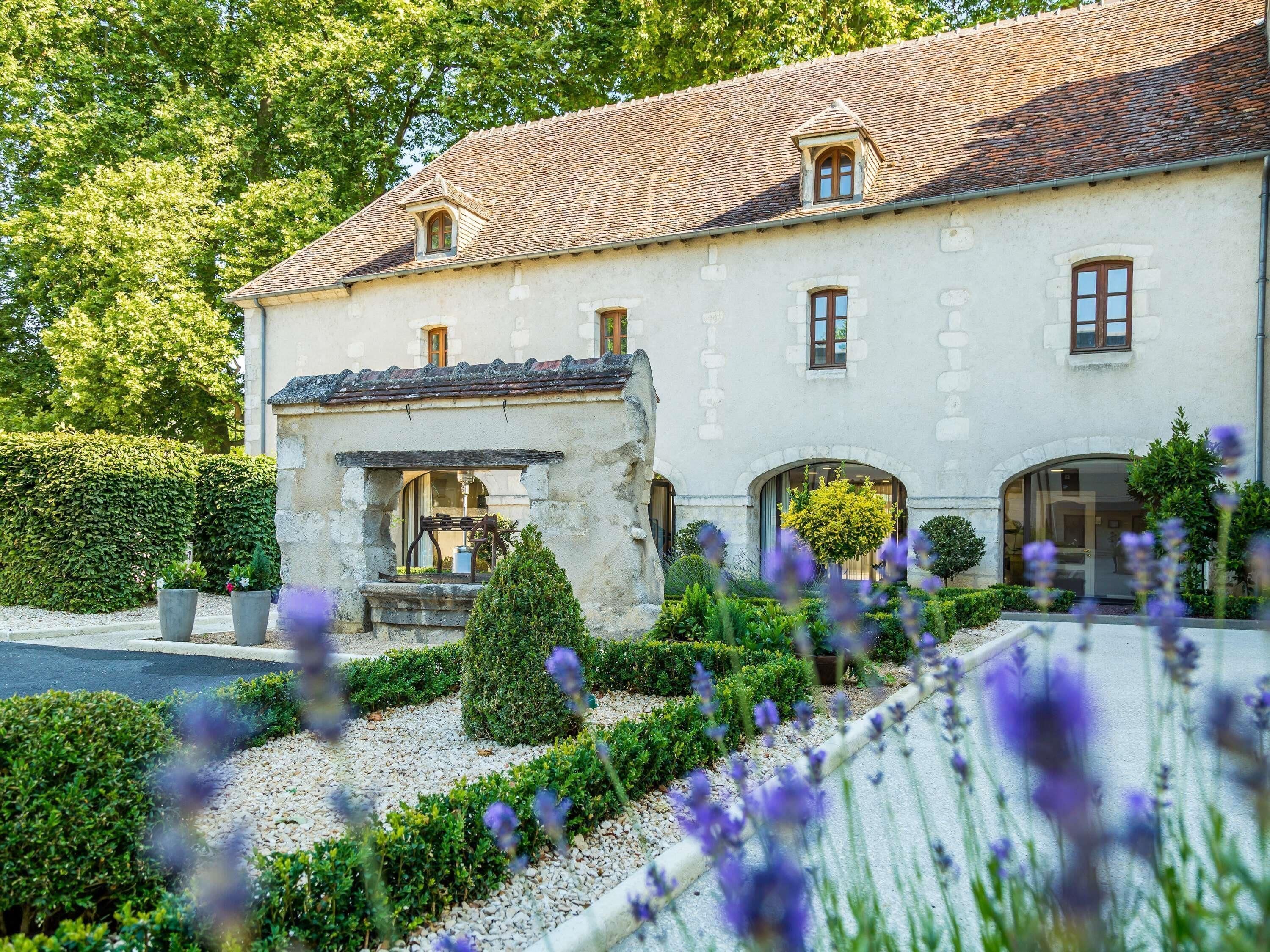 Un grand bâtiment historique, doté d’un toit recouvert de tuiles, de nombreuses fenêtres et d’un jardin bien entretenu où poussent diverses plantes et fleurs.