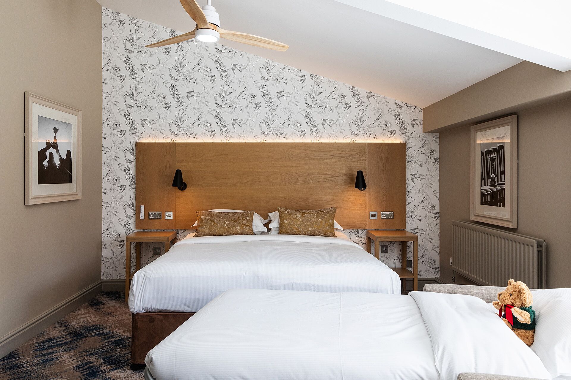 A hotel room with two beds, a wooden headboard, a ceiling fan, and floral wallpaper.