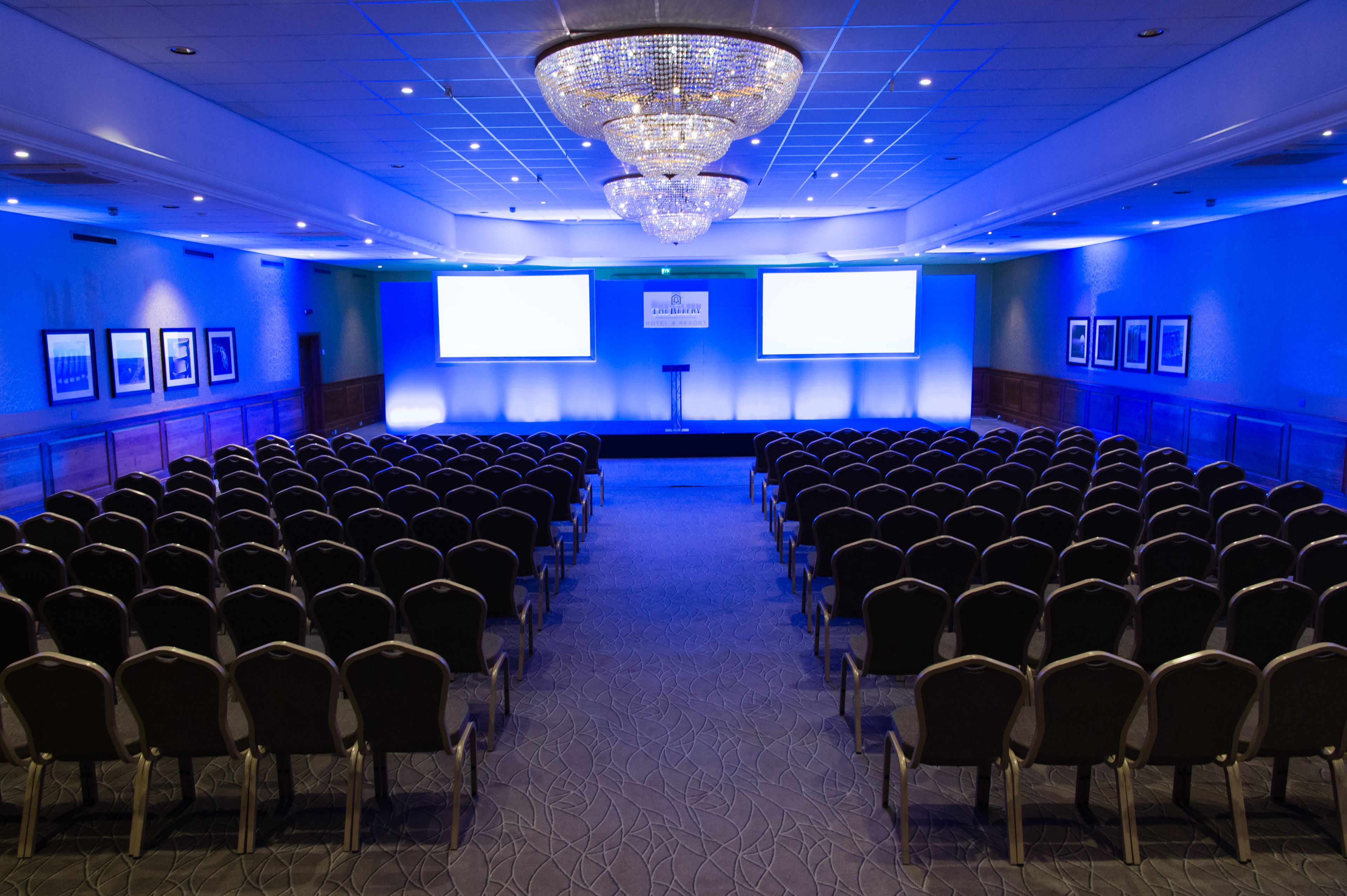 A conference room with rows of chairs, a large chandelier, and two screens on the back wall.
