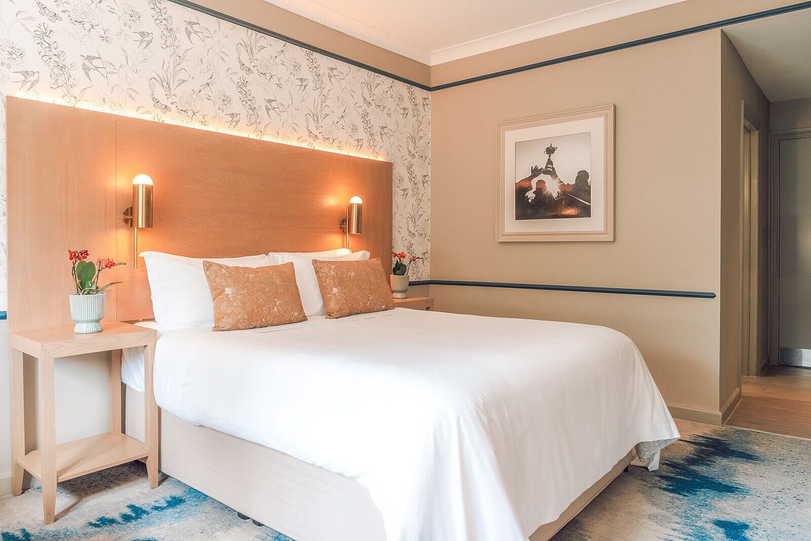 A neatly made bed with white linens and two decorative pillows, a bedside table with a vase of flowers, and a framed picture on the wall.