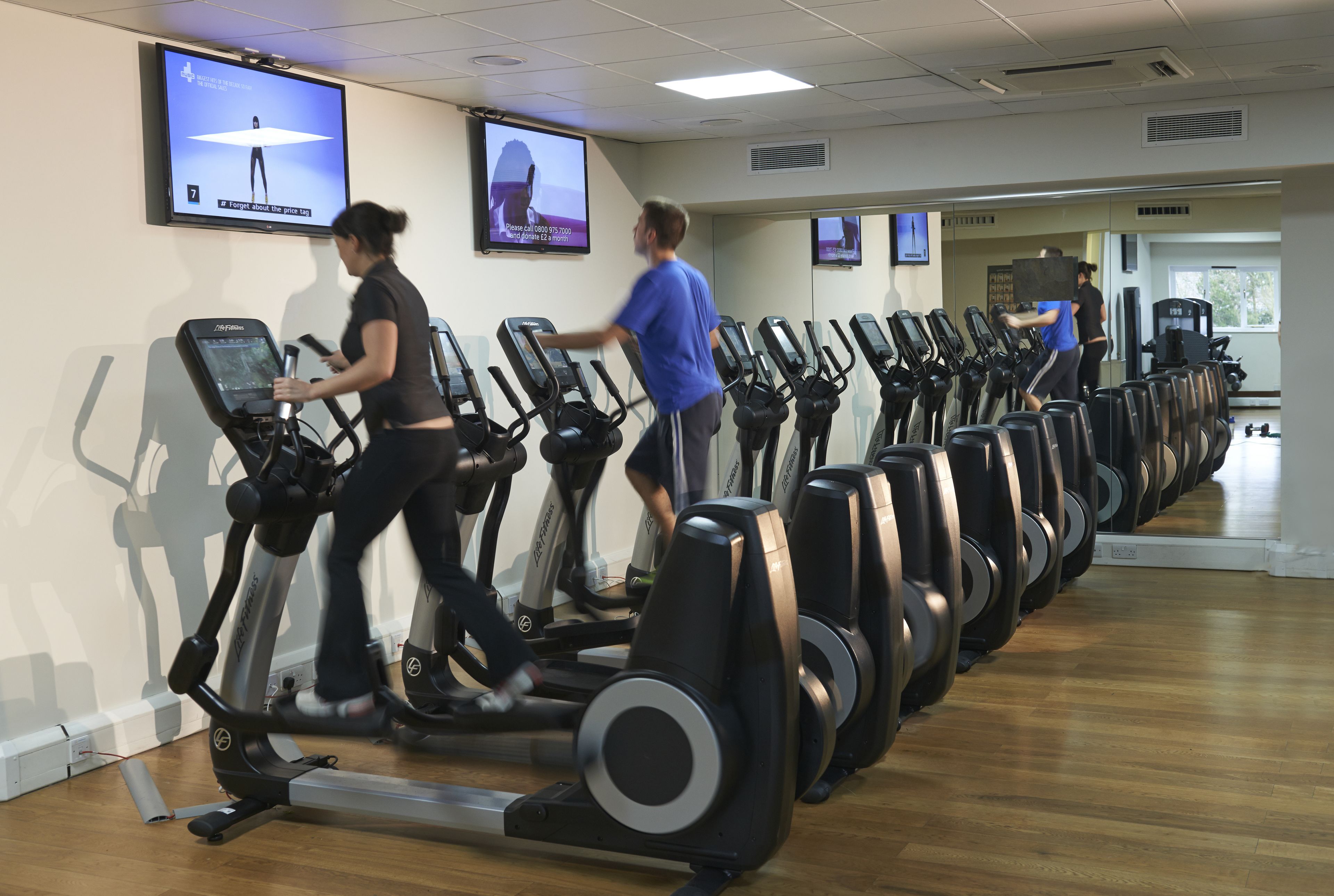 A gym with multiple treadmills lined up against the wall, a large mirror, and a television screen displaying a workout video.
