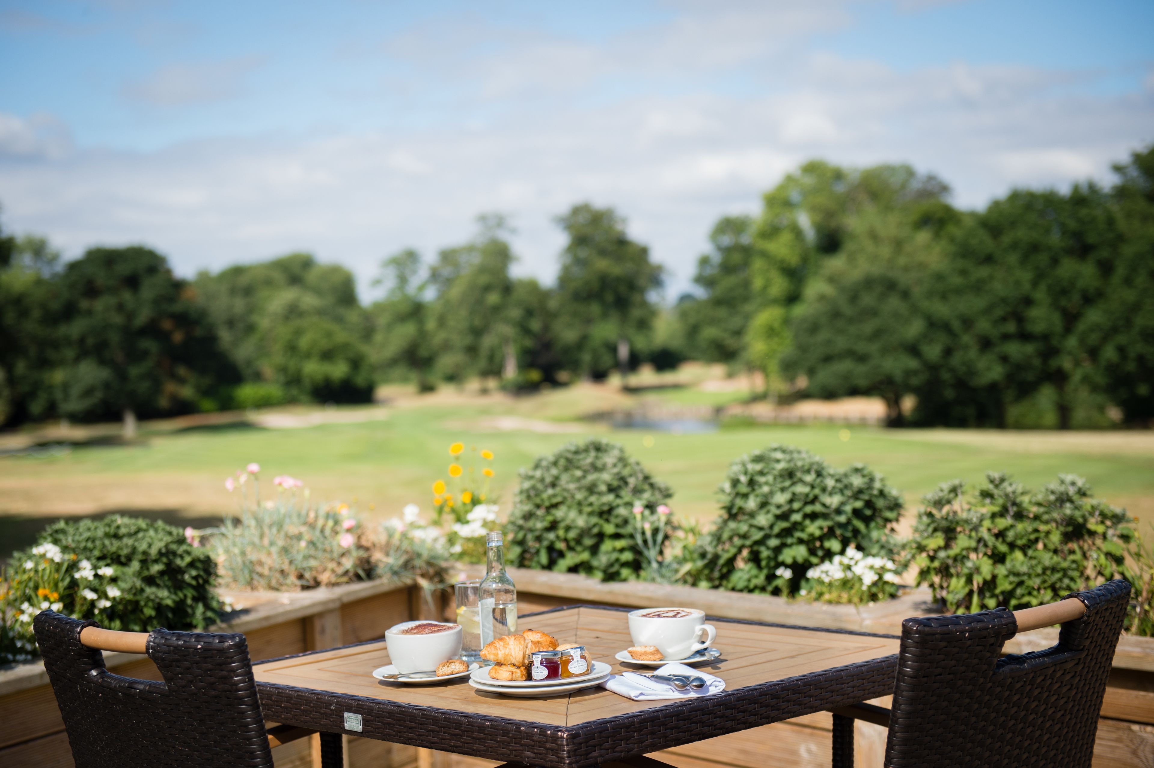A dining table with a croissant, coffee, and a bottle, set against a backdrop of a golf course and greenery.