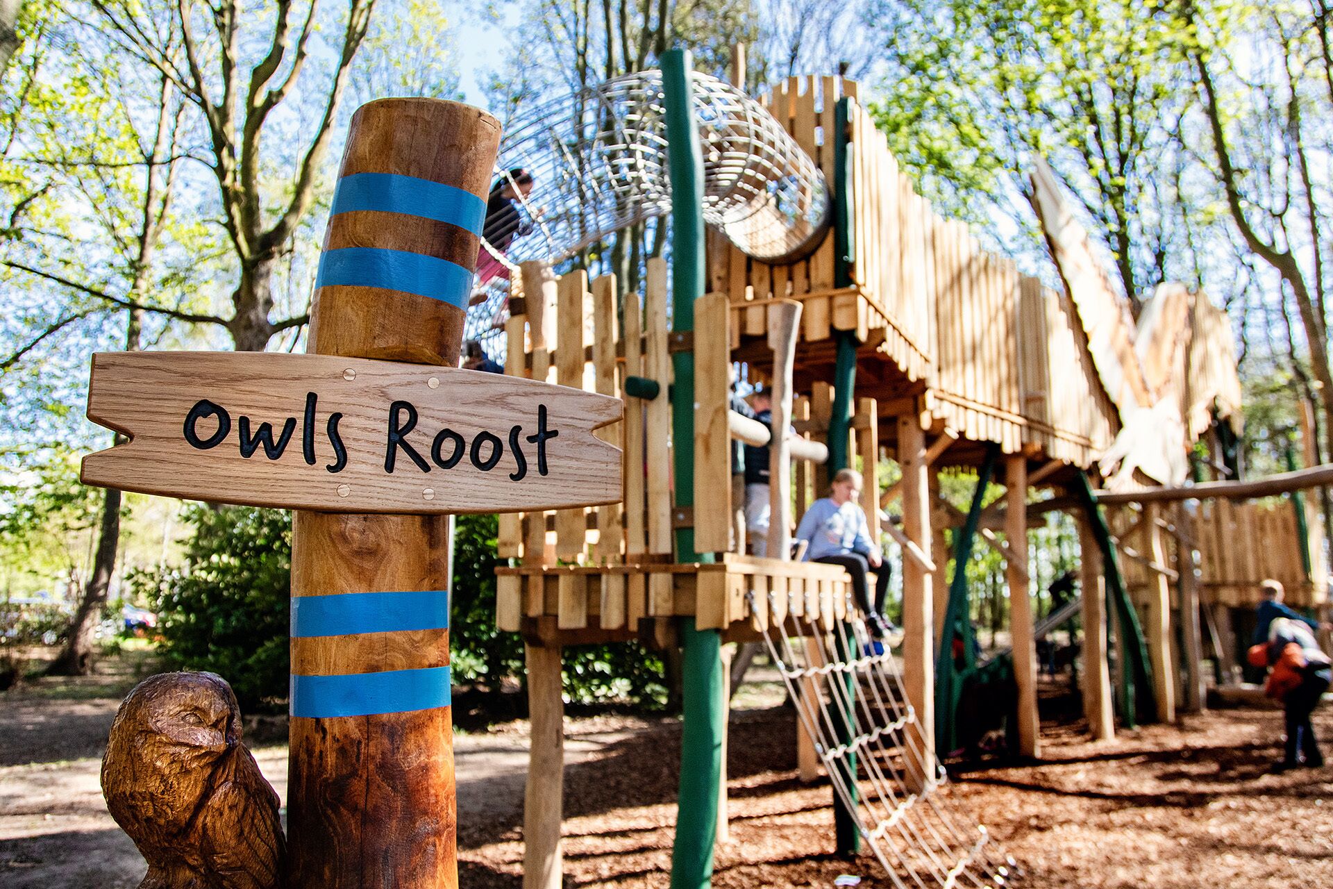 A wooden signpost with 'Owls Roost' written on it, part of a wooden play structure in a park.