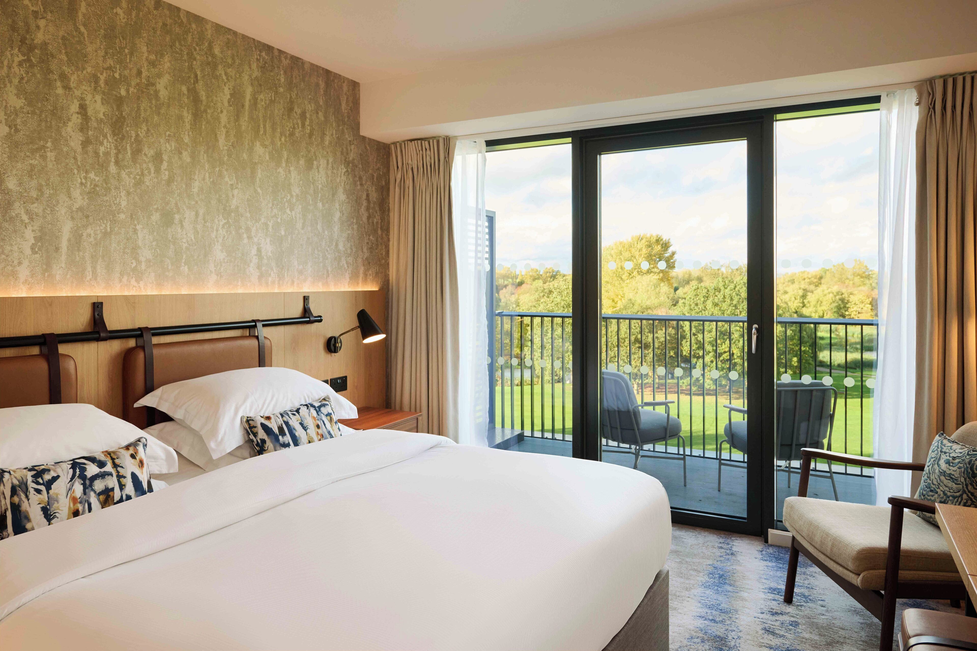 A hotel room with a large bed, a desk, a chair, and a balcony with a view of the greenery.