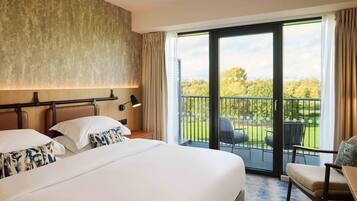 A hotel room with a large bed, a desk, a chair, and a balcony with a view of the greenery.