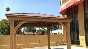 A wooden pergola structure with a metal roof, set against a backdrop of a building with a yellow facade and large windows. - Riverside Inn & Suites (Riverside)