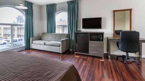 A hotel room with a bed, a sofa, a desk with a computer, a TV, and a refrigerator. - Riverside Inn & Suites (Riverside)