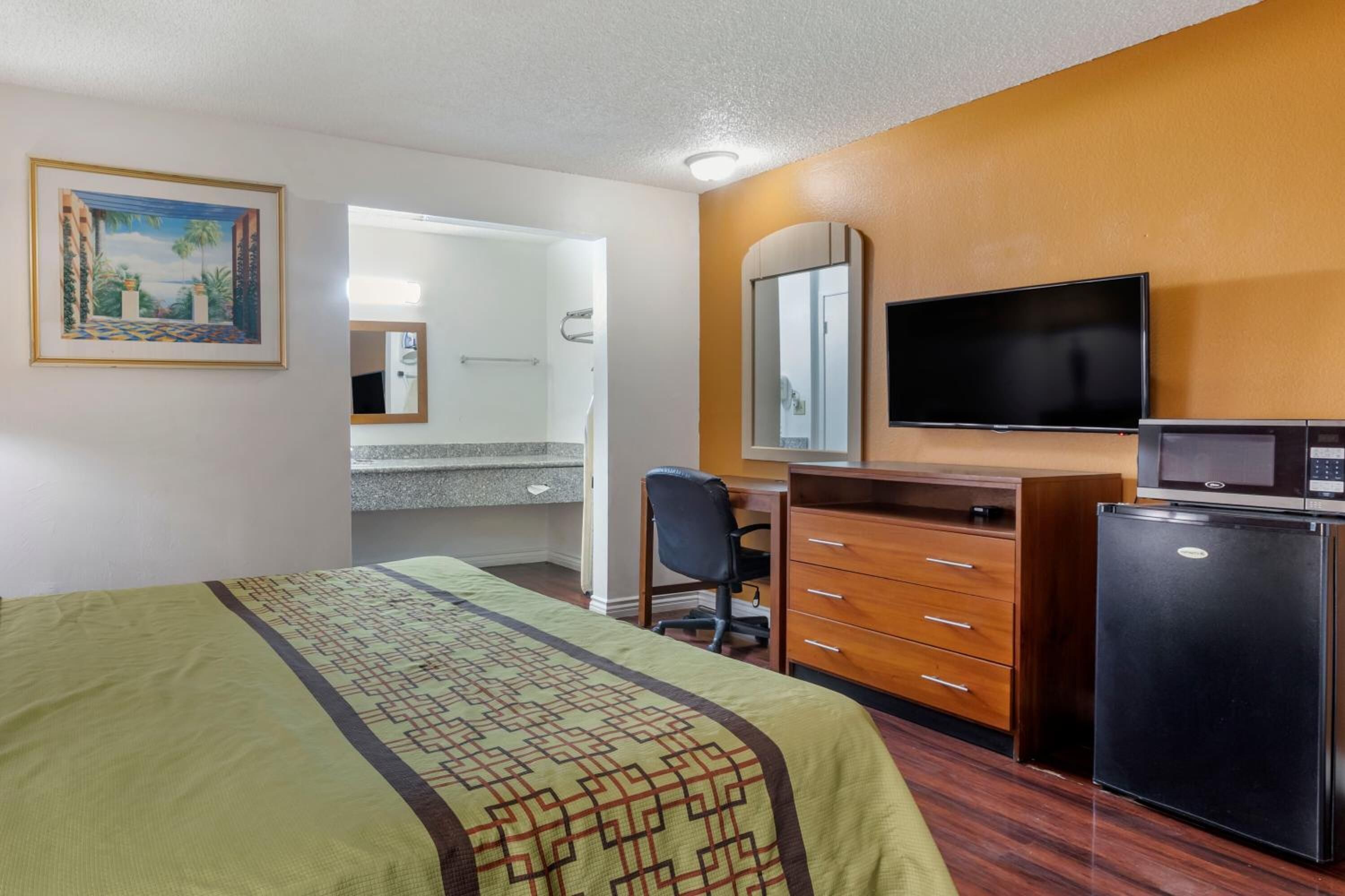 Quarto de hotel com cama, escrivaninha com cadeira, televisão, frigorífico e casa de banho visível por uma porta aberta.