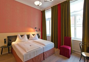 Quarto de hotel com uma cama grande, uma cadeira, uma mesinha e uma janela com cortinas.