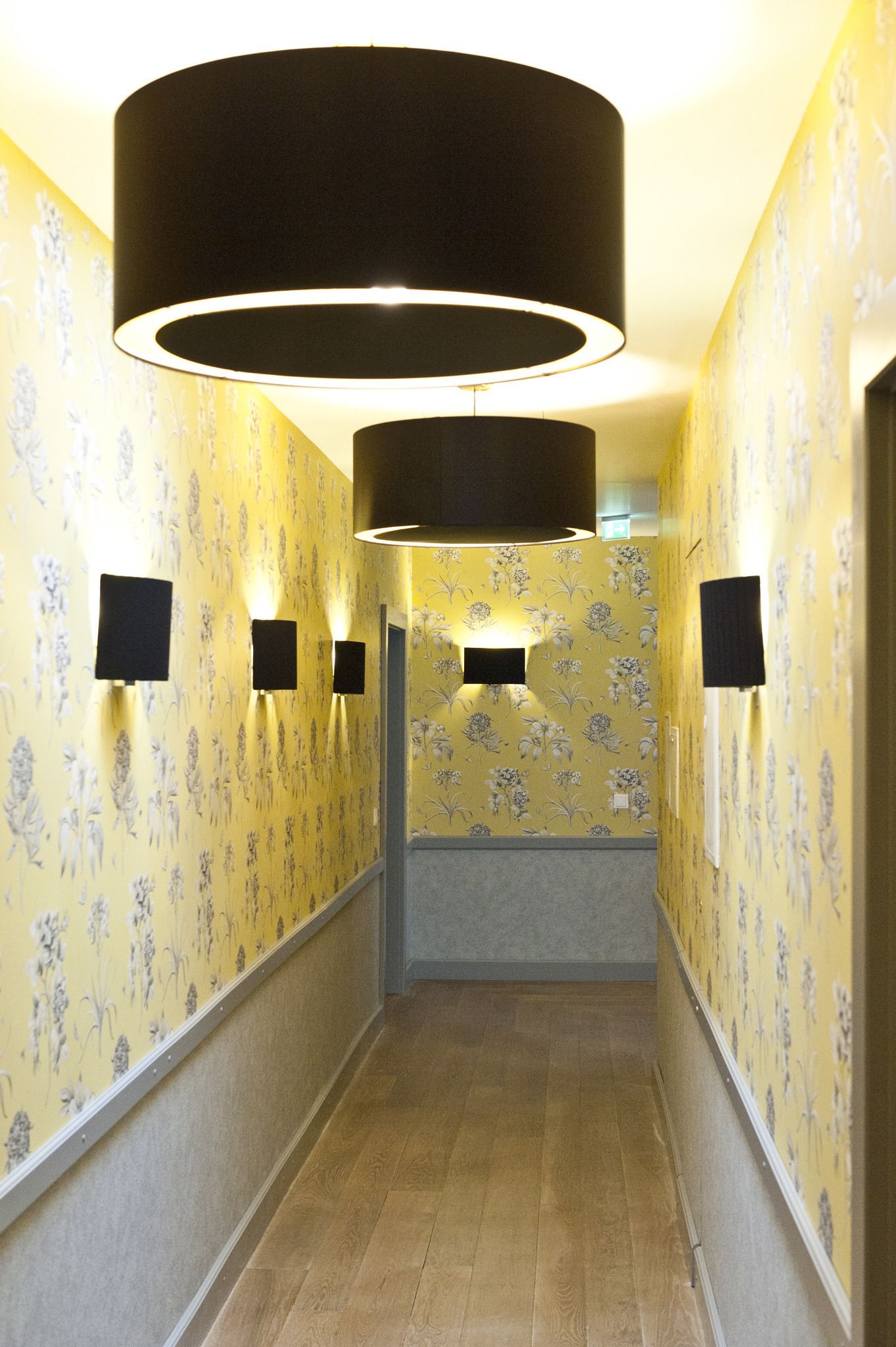 Un couloir avec un papier peint à motifs, des appliques murales noires et un grand luminaire suspendu noir.
