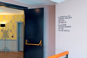 Quarto de hotel com porta preta, corrimão de madeira e parede com indicação do andar 6.
