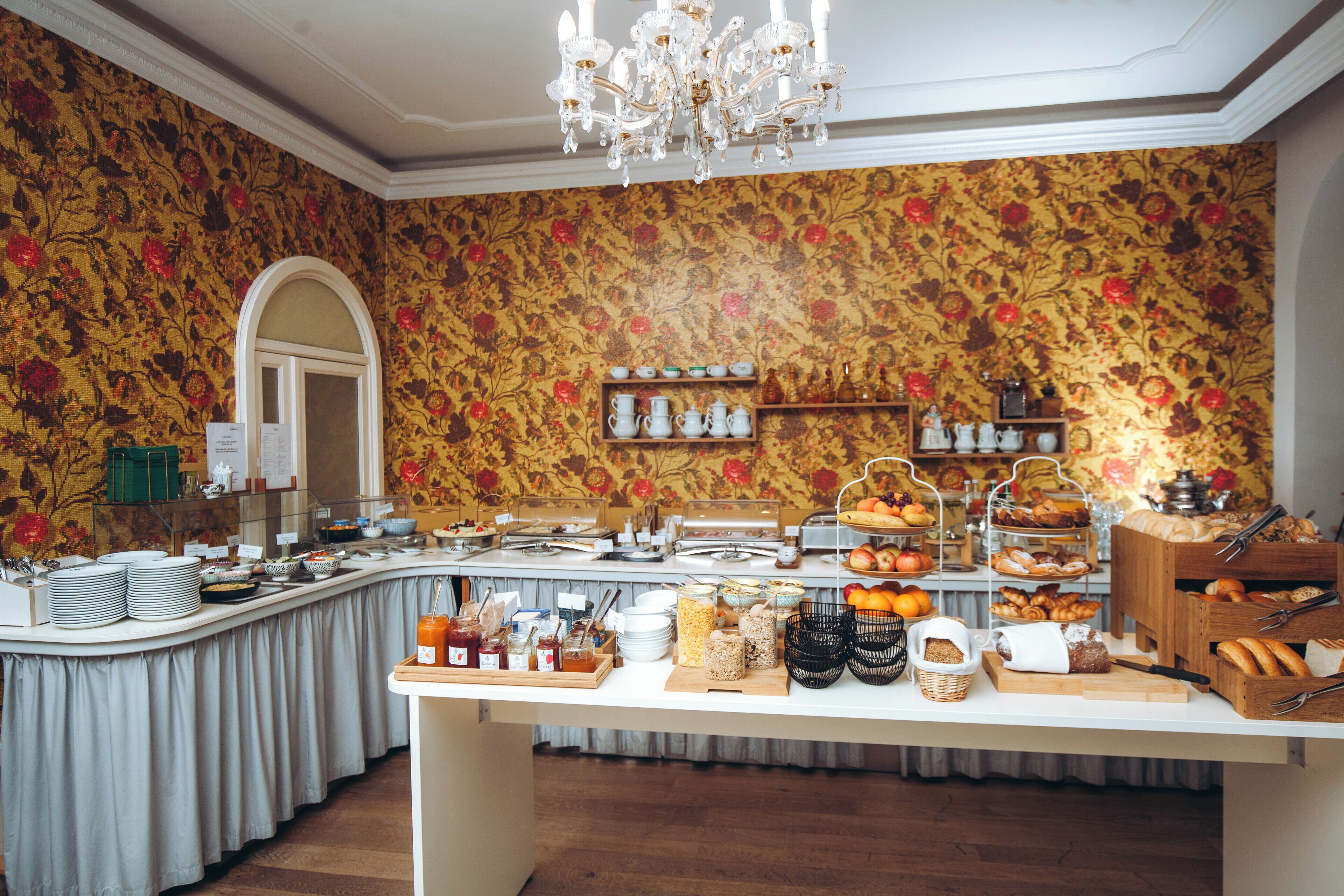 A buffet setup with various food items and utensils, a chandelier, and a floral wallpaper background.