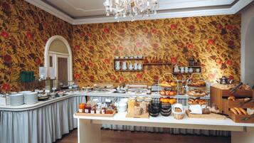 A buffet setup with various food items and utensils, a chandelier, and a floral wallpaper background.