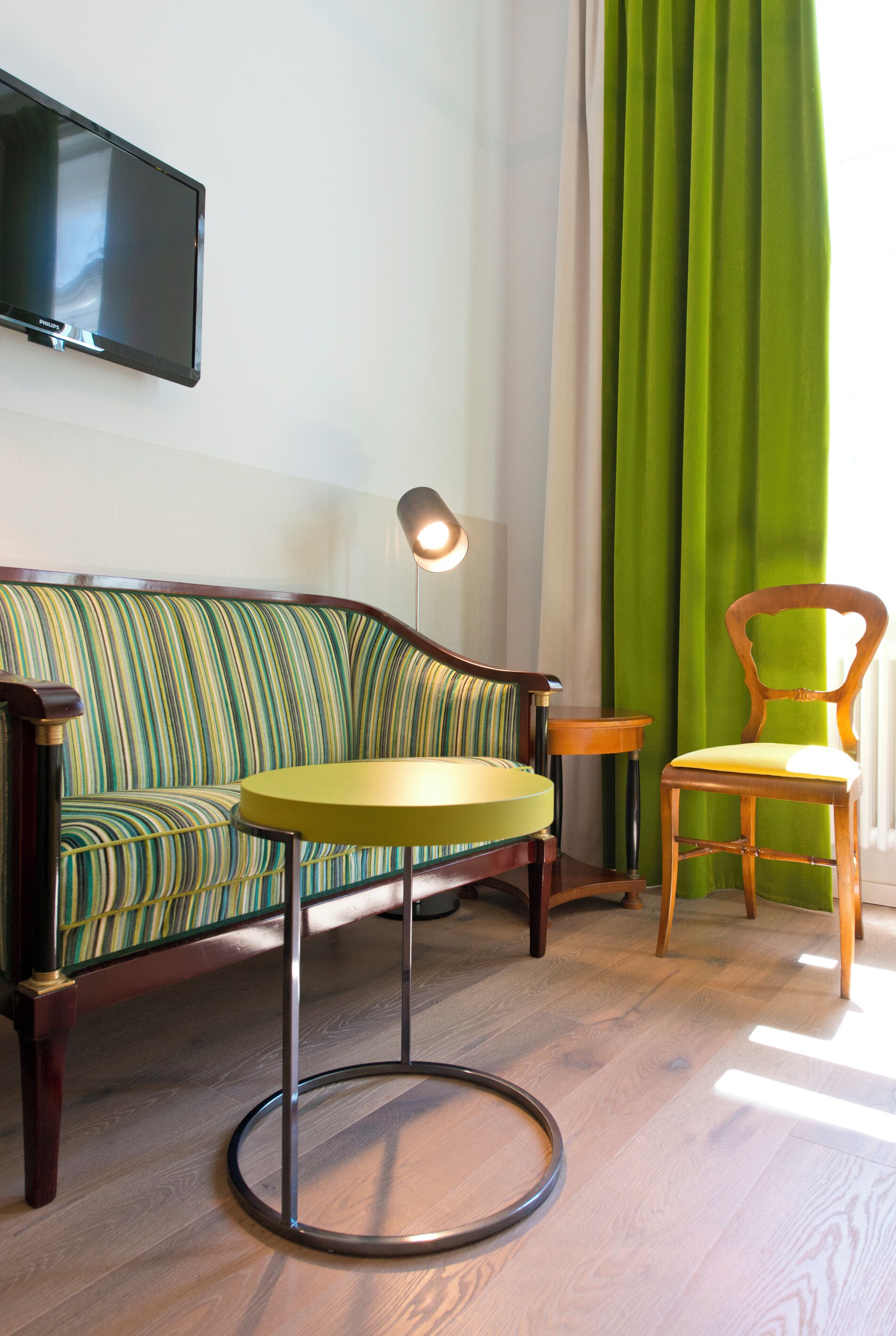 A room with a striped sofa, a round table, a wooden chair, a lamp, and a television on the wall.