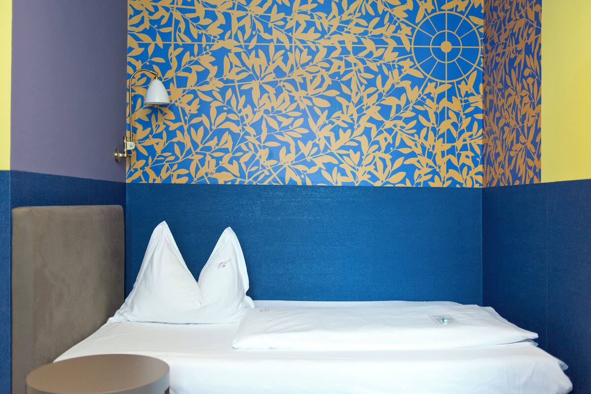 Una habitación de hotel con una cama, un almohadón, una mesita de noche y una pared con estampado azul y amarillo.