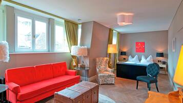 Une chambre d’hôtel moderne avec un canapé rouge, un fauteuil à motifs floraux, une commode en bois et un lit avec des oreillers blancs.