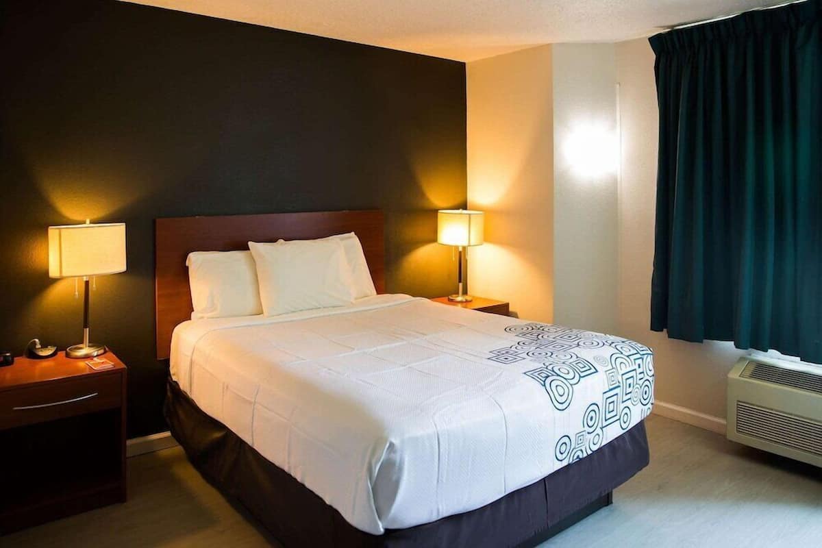 A hotel room with a large bed, two bedside tables with lamps, a window with curtains, and a wall-mounted air conditioner.
