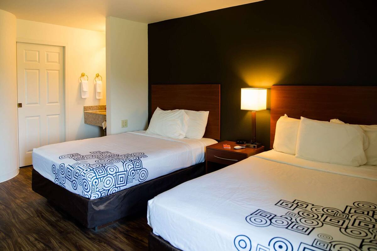 A hotel room with two beds, a wooden headboard, a nightstand, a lamp, and a bathroom with a white door and towel rack.