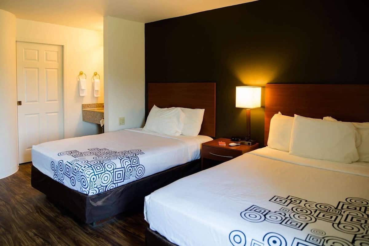 A hotel room with two beds, a wooden floor, a dark wall, and a nightstand with a lamp.