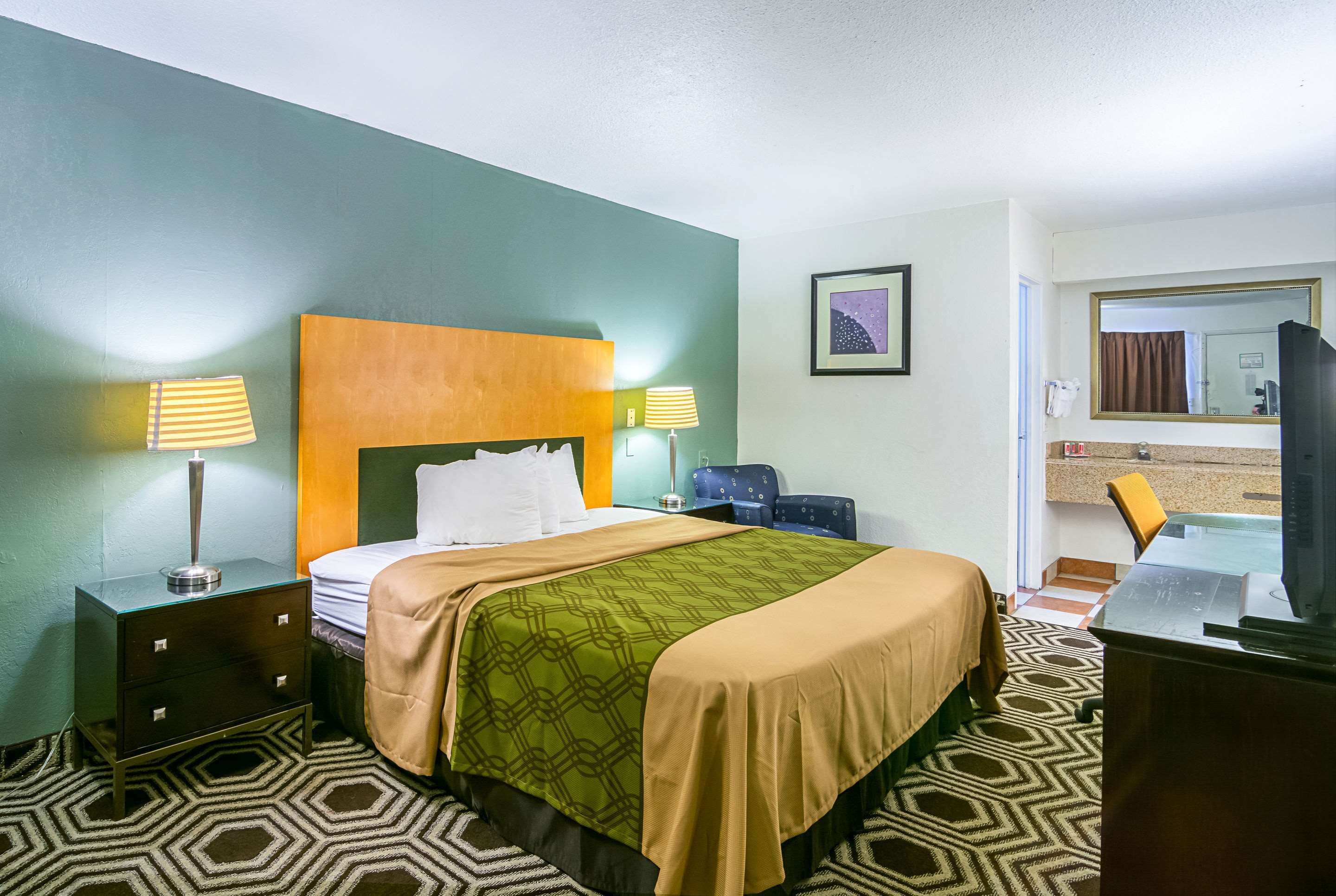 A hotel room with a bed, bedside tables, a desk, a chair, a mirror, and a bathroom visible through an open door.