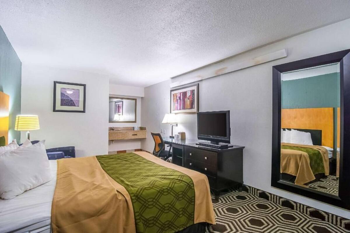 Een hotelkamer met een bed, een bureau met een televisie, een stoel, een dressoir en een grote spiegel.