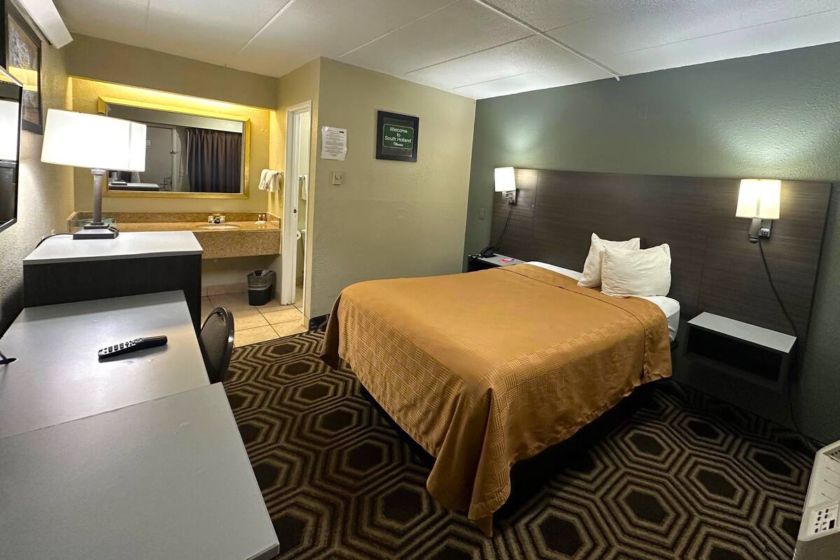 Een hotelkamer met een bed, nachtkastje, lamp en een klein bureau met een afstandsbediening.