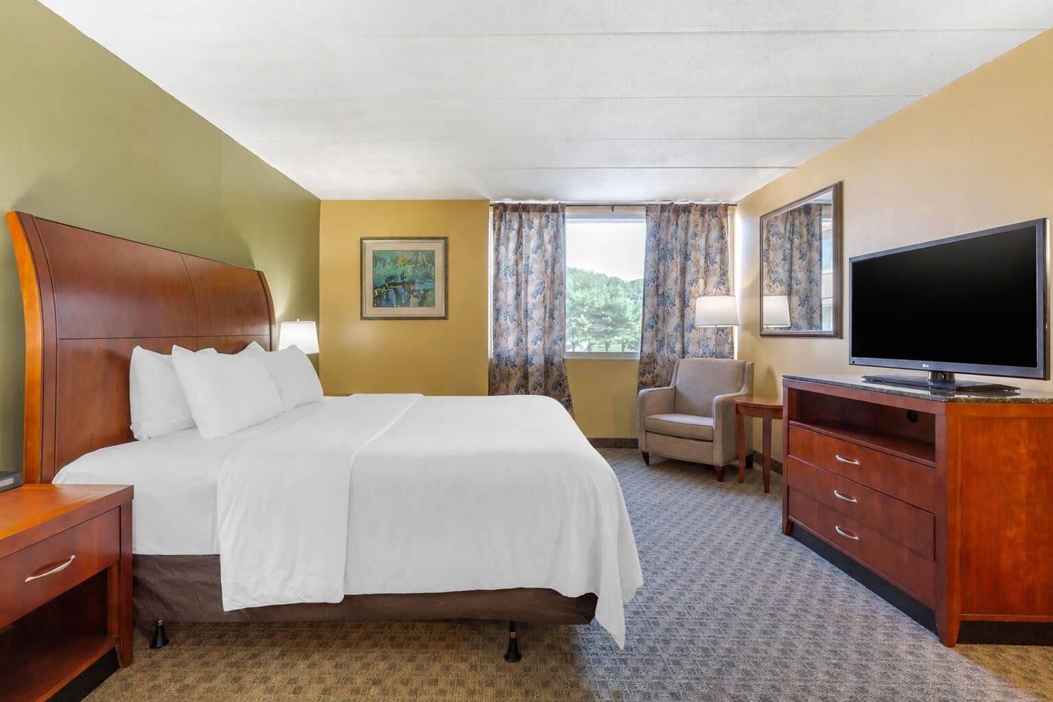A hotel room with a bed, a TV, a chair, a desk, and a painting on the wall.