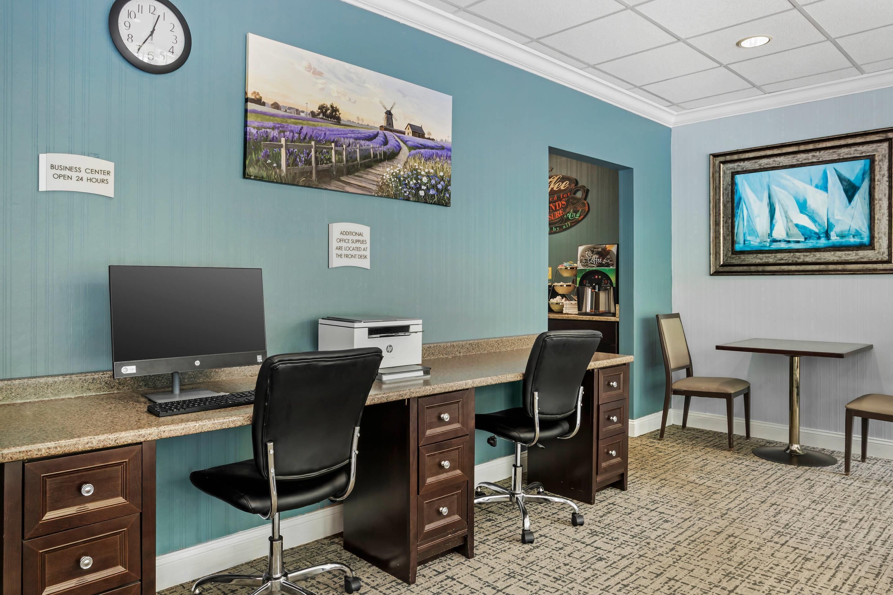 A business center with a desktop computer, two chairs, a clock, and a painting on the wall.