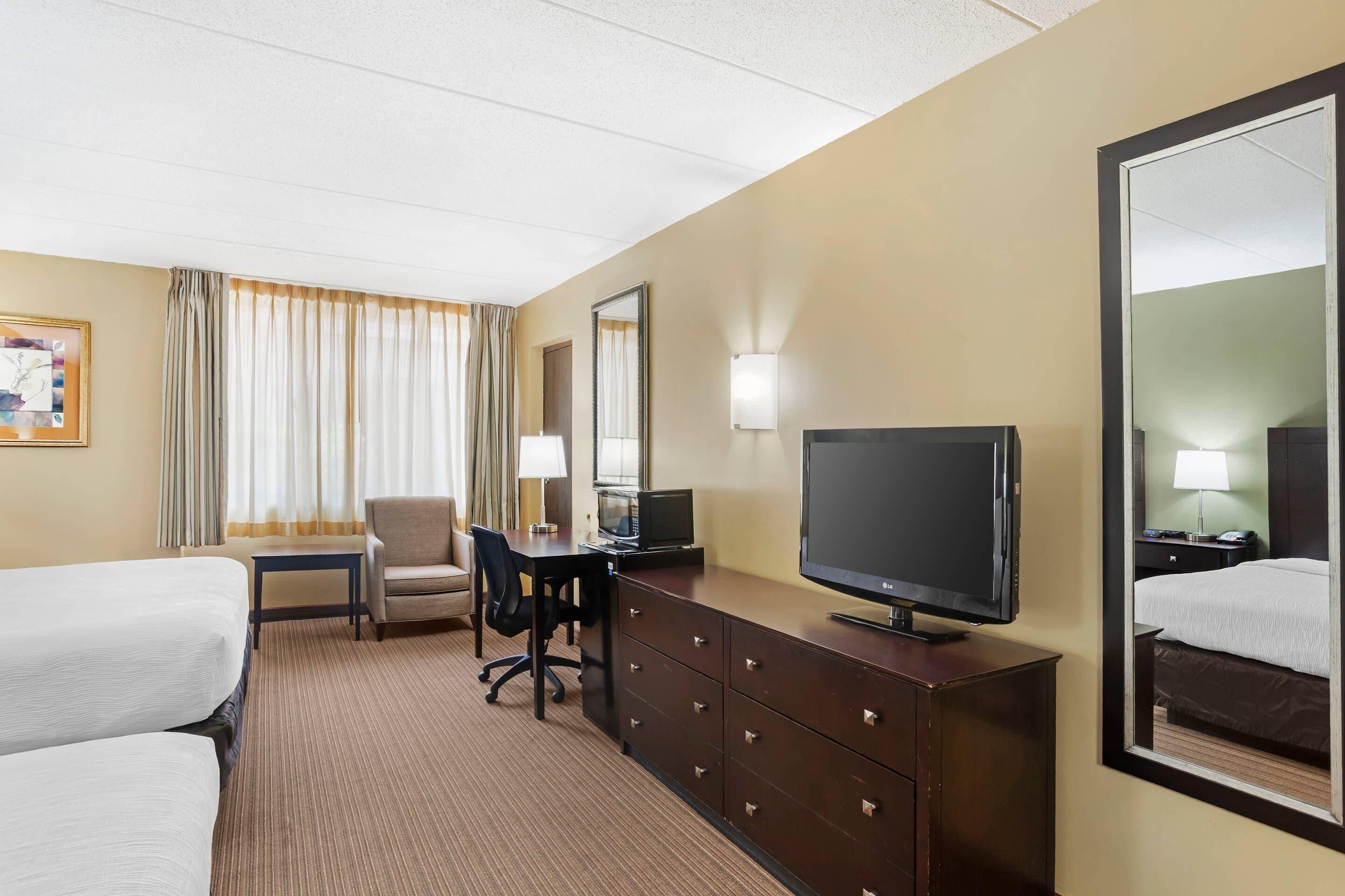 Habitación de hotel con cama, escritorio, computadora, televisor y un espejo grande.