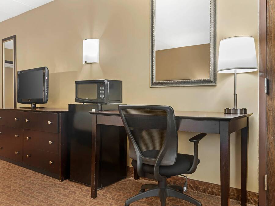 A hotel room with a desk, a television, a microwave, a chair, a dresser, and a mirror.