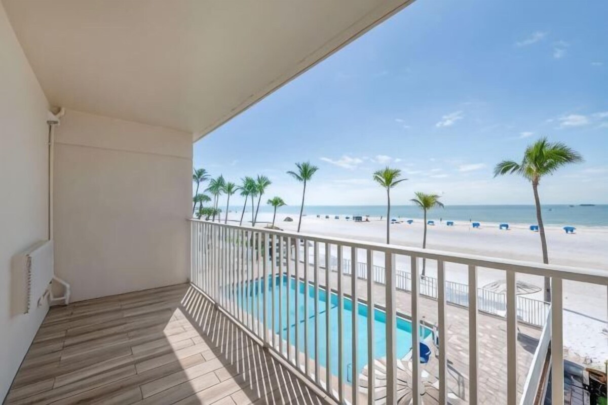A balcony with a view of a beach, a pool, and palm trees.