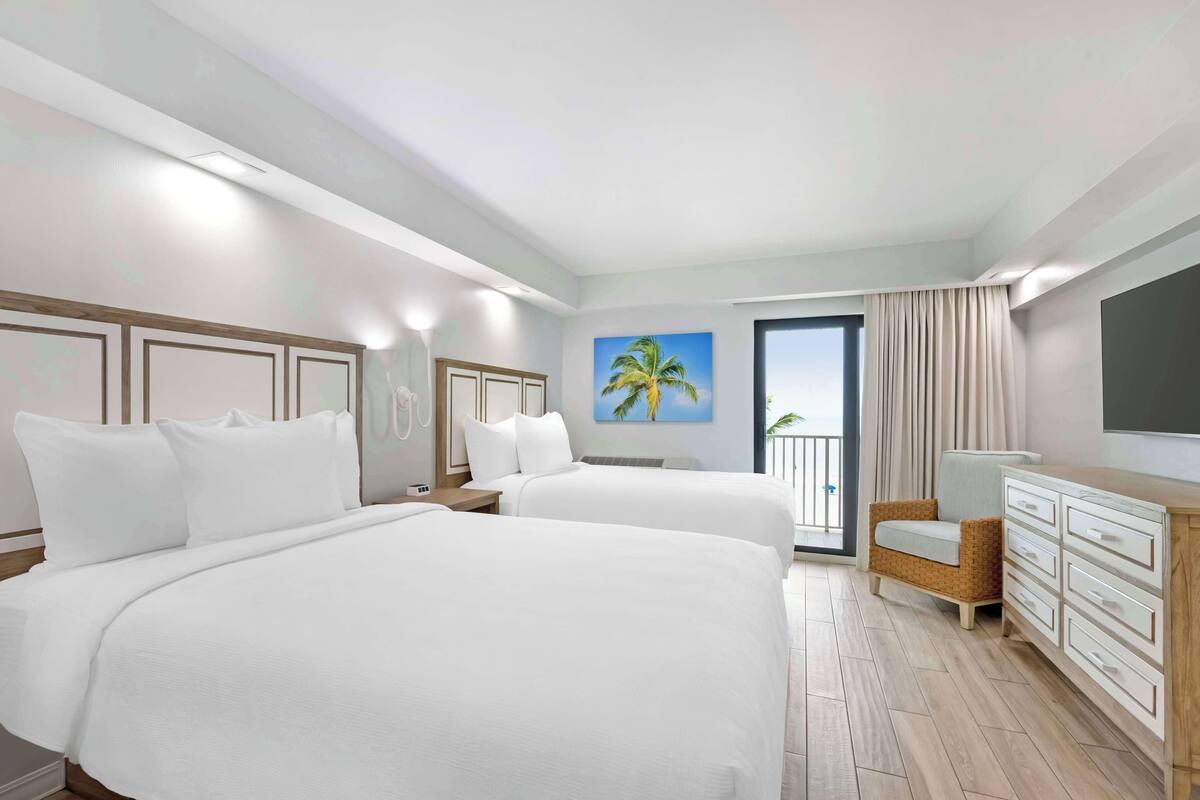 Habitación de hotel con dos camas, un aparador de madera, una silla y un cuadro de una palmera.
