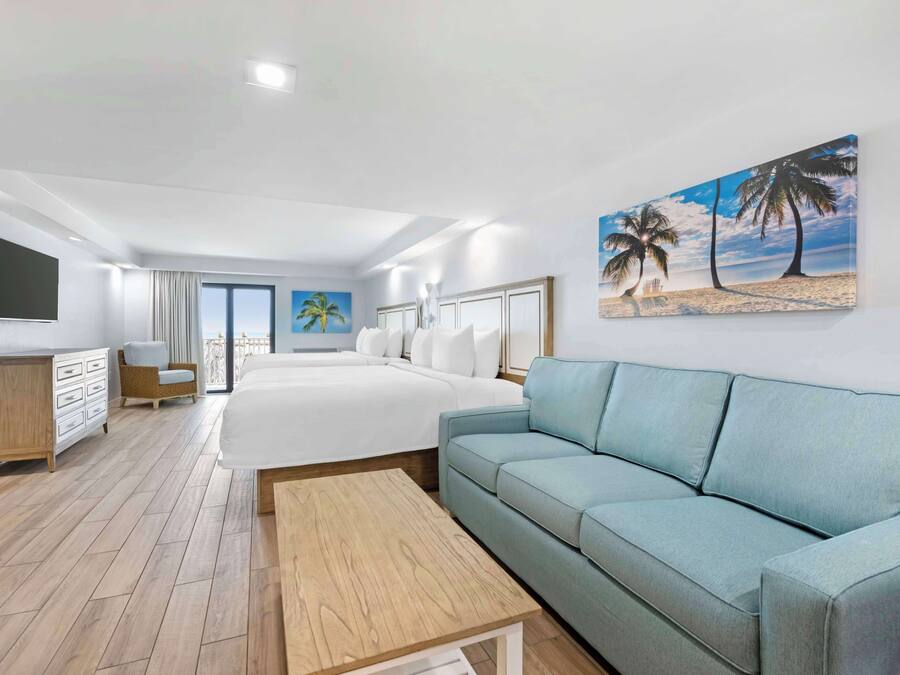 A modern hotel room with a large bed, a light blue sofa, a wooden coffee table, a TV mounted on the wall, and a painting of a beach scene.