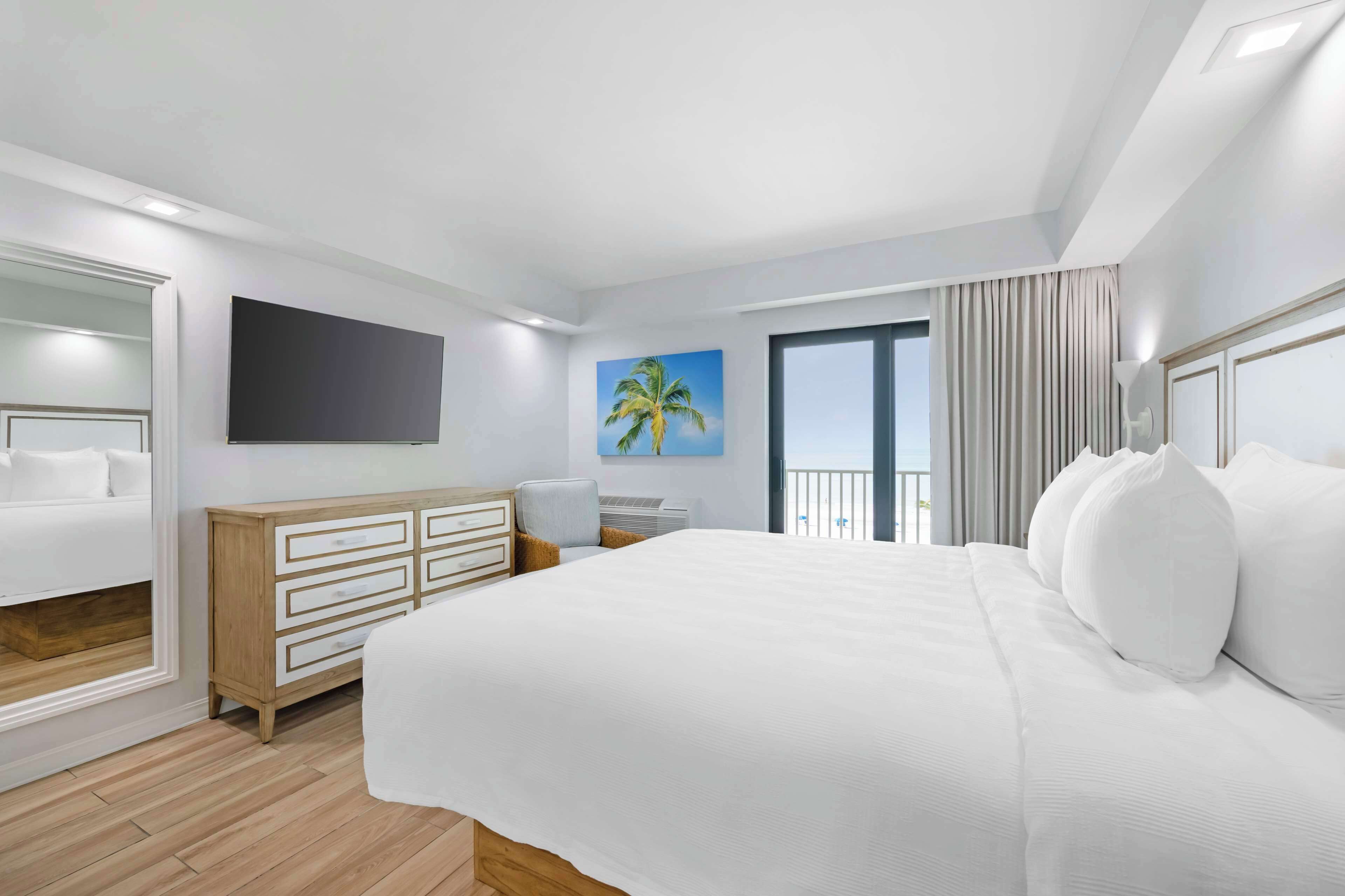 A modern hotel room with a large bed, a dresser, a flat-screen TV, a painting of a palm tree, and a balcony with curtains.