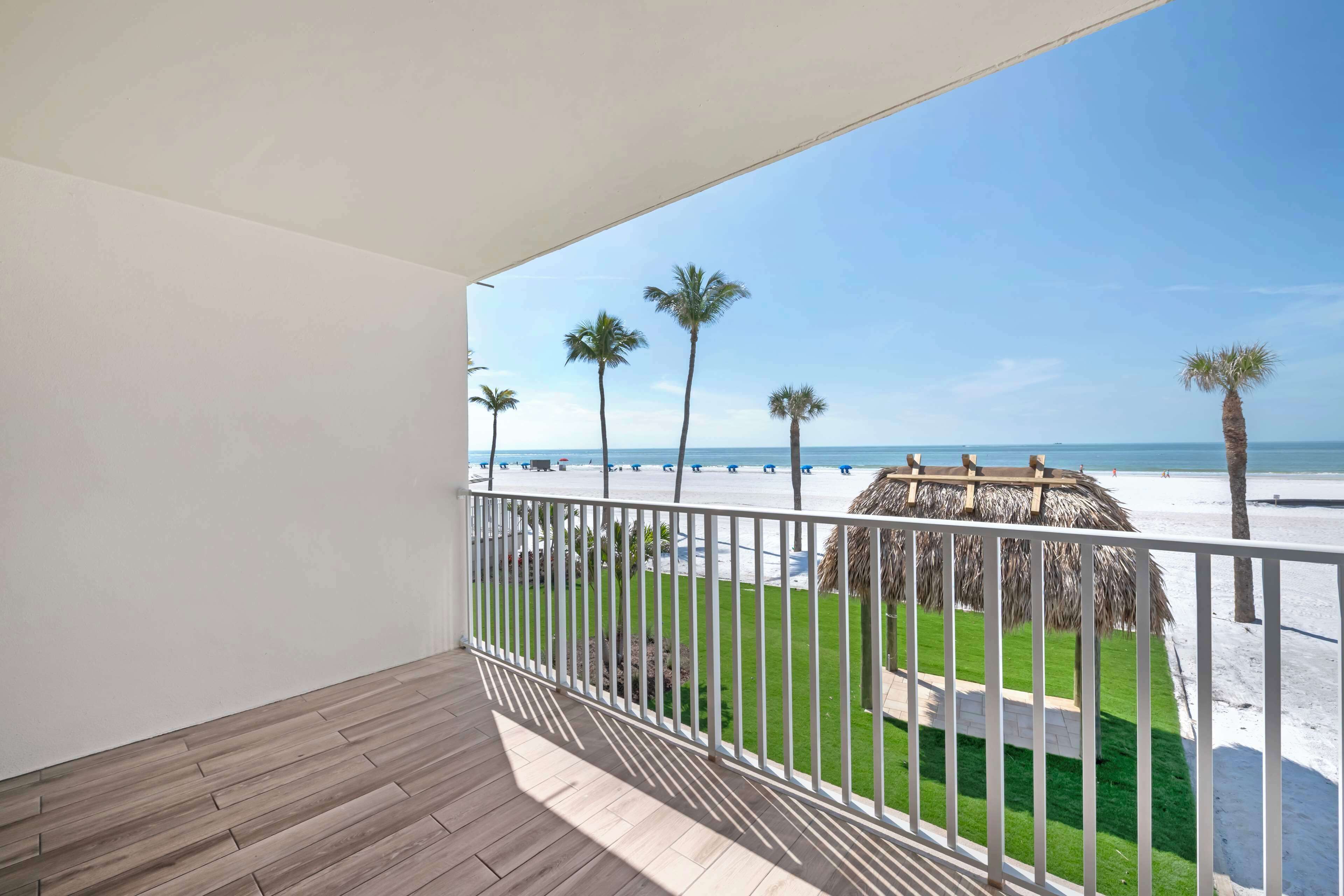 A balcony with a wooden floor and white railing, overlooking a beach with palm trees and a thatched structure.