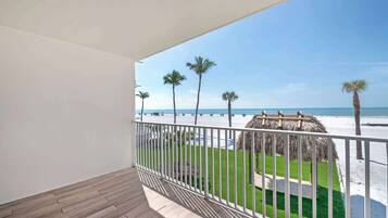 A balcony with a wooden floor and white railing, overlooking a beach with palm trees and a thatched structure.