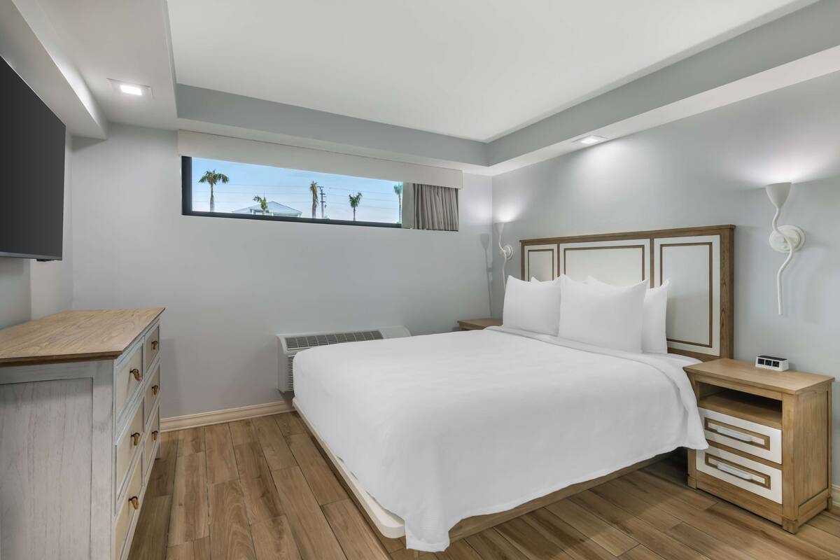 Un dormitorio moderno con una cama grande, una mesita de noche, un armario y una ventana con vistas a palmeras.